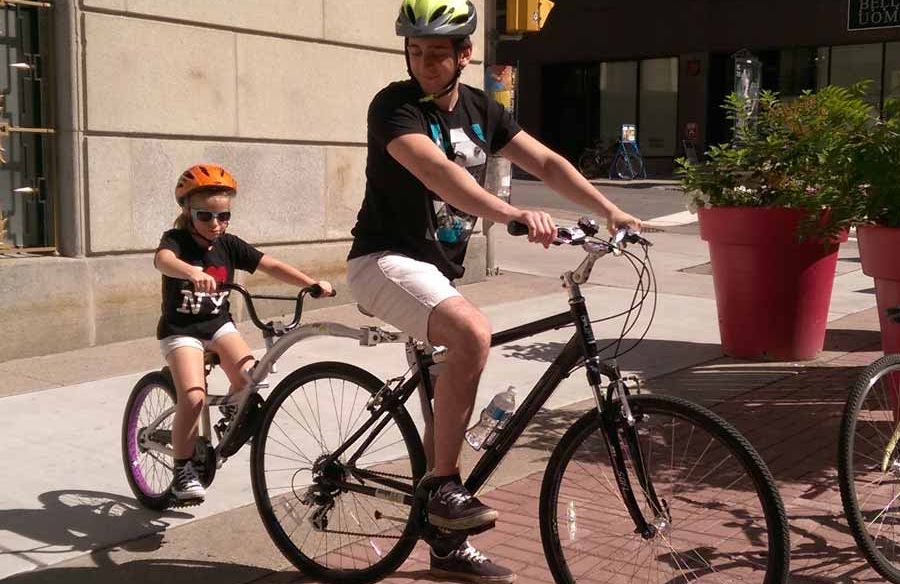 A parent is biking with the kid riding the rental trail-a-bike attached to his bike on Sparks St., Ottawa
