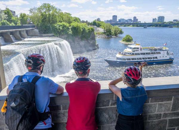 Guests are taking an Escape bike & boat tour and stop at Rideau Falls Ottawa landmark to watch a boat cruise