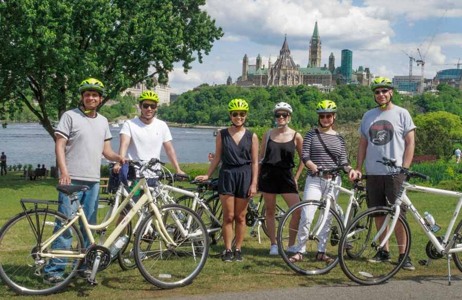 Guests are taking a group picture with a view of Ottawa River and parliament building during a scheduled Ottawa bike tour with Escape
