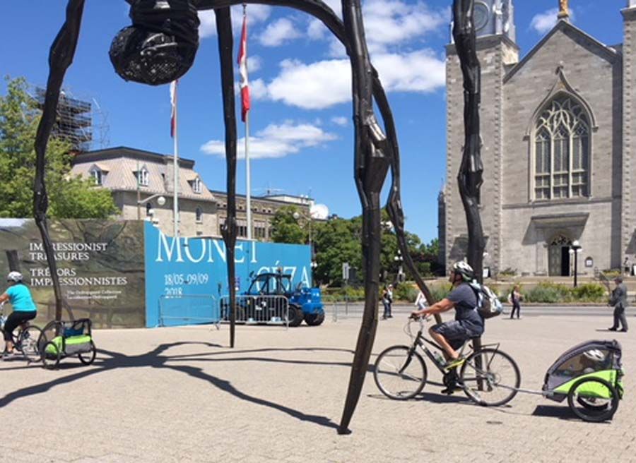 Parents biking with kids sitting on Escape rental child trailers, visiting Ottawa landmark, Maman spider sculpture 