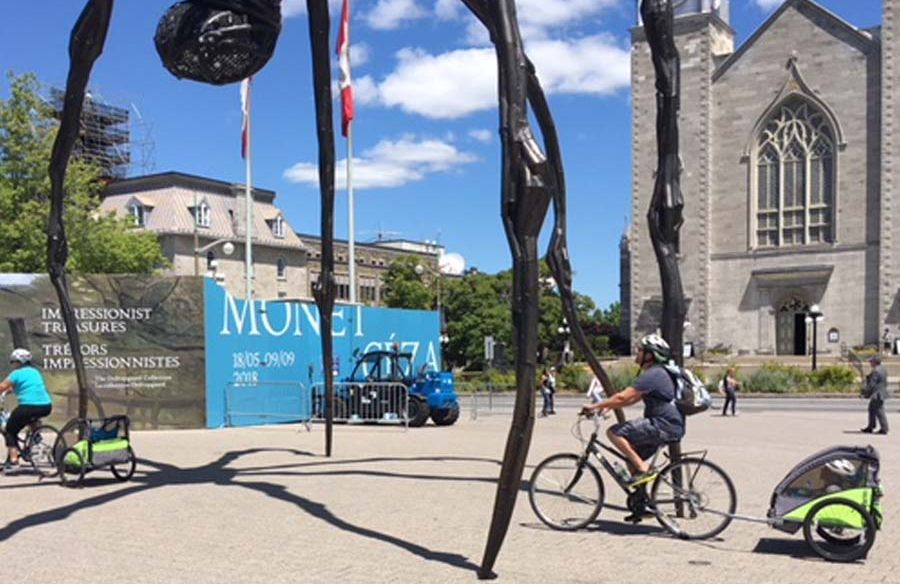 Parent biking with kids sitting on Escape rental child trailers, visiting Ottawa landmark, Maman spider sculpture 