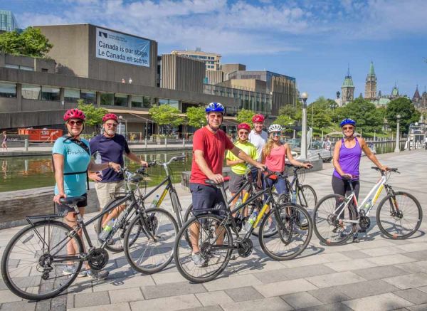 Guest are biking on Rideau Canal Pathway with Escape Bike Tour with the view of National Art Centre and Parliament Building