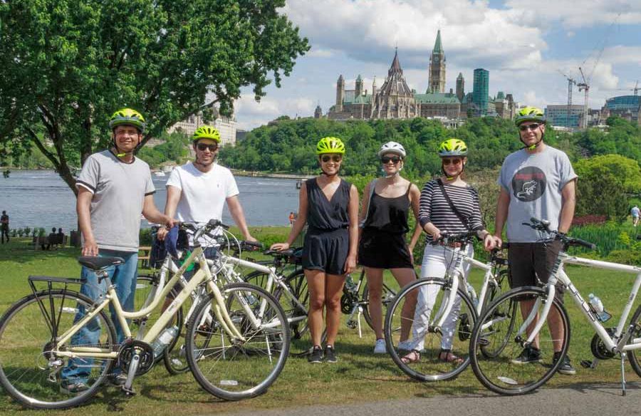 Guests are taking a group picture with a view of Ottawa River and parliament building during a scheduled Ottawa bike tour with Escape 